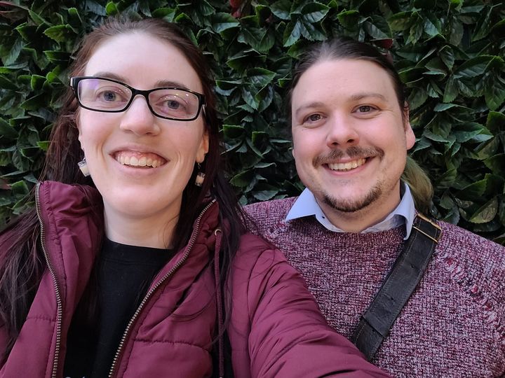 [Image description: Two pale white humans standing against a leafy backdrop. Emma is on the left and has glasses, brown hair, a red puffy jacket and bubble tea earrings. To the right stands *wild husband. Temporarily domesticated until someone offers him cheese.* He has facial hair and wears a red jumper. They are both smiling at the camera. End description.]